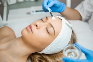 Beautician's hands in blue protective gloves applying a transparent moisturizing peeling mask to a woman's facial skin. Chemical Peel Beauty Treatment